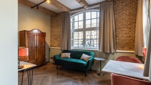 A room with a wooden floor, a green sofa, a wooden cabinet, a small round table, and a window with curtains. - Studio Urbain (Alkmaar)