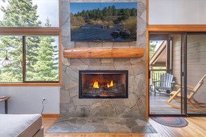A stone fireplace with a fire, a view of a river, and a wooden deck with Adirondack chairs. - Updated Pet-Friendly Trailside Retreat (Truckee)