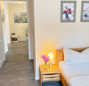 Une chambre d’hôtel avec un plancher en bois, un lit avec une literie à rayures, une table de chevet avec une lampe et des fleurs, et un couloir menant à une autre pièce.