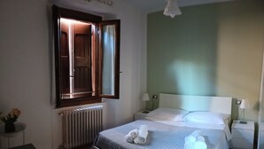 Une chambre d’hôtel avec un lit, une table de chevet, une lampe, une fenêtre avec des rideaux et un radiateur.