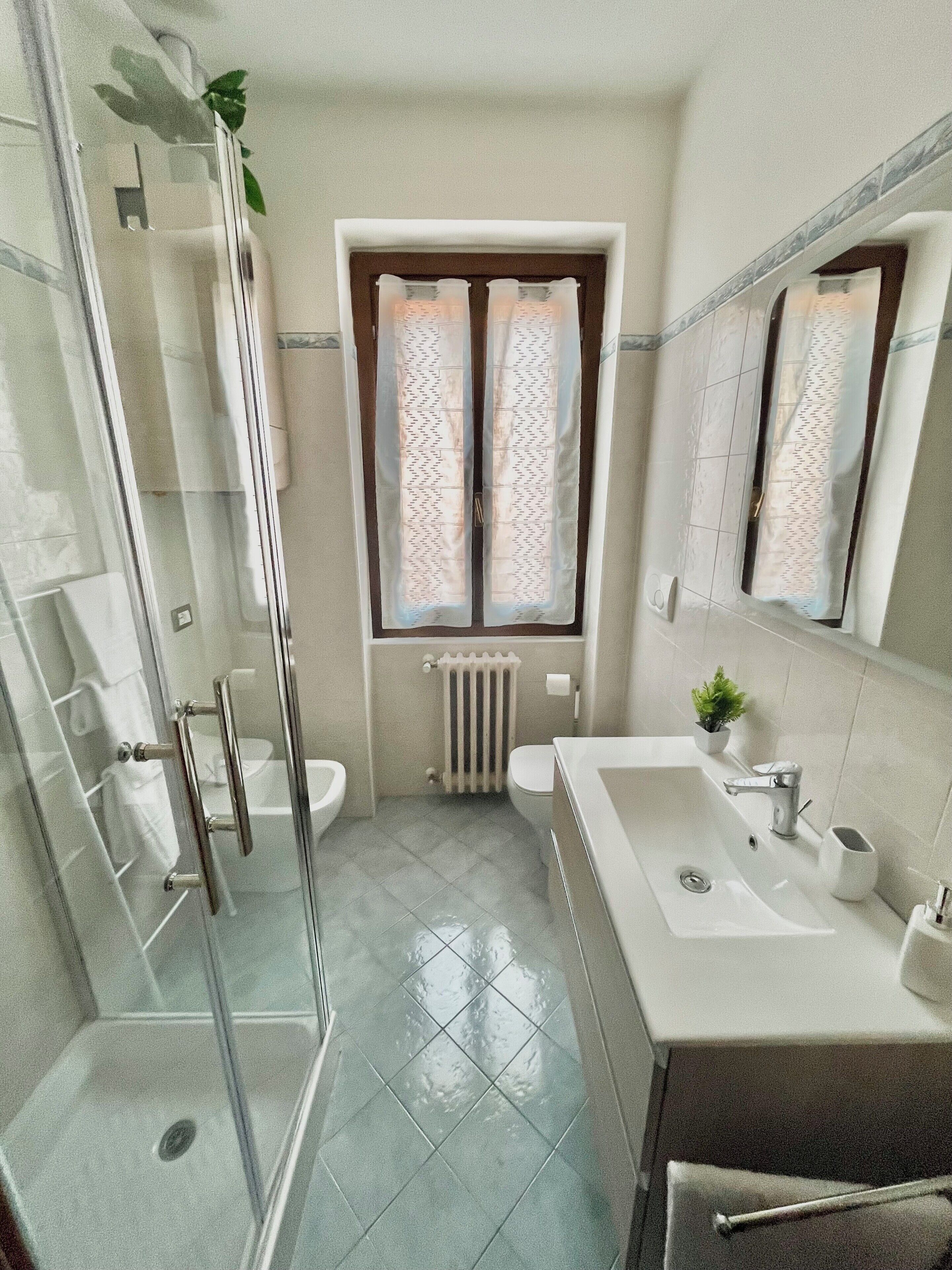 A bathroom with a shower, toilet, sink, and window with curtains.. The House of Agnes