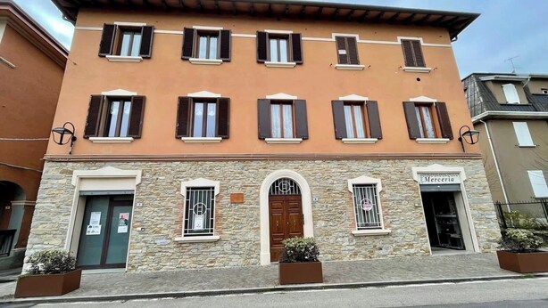 Un bâtiment à deux étages, doté d’une façade en pierre, de nombreuses fenêtres avec volets et d’une enseigne indiquant « Merceria ».