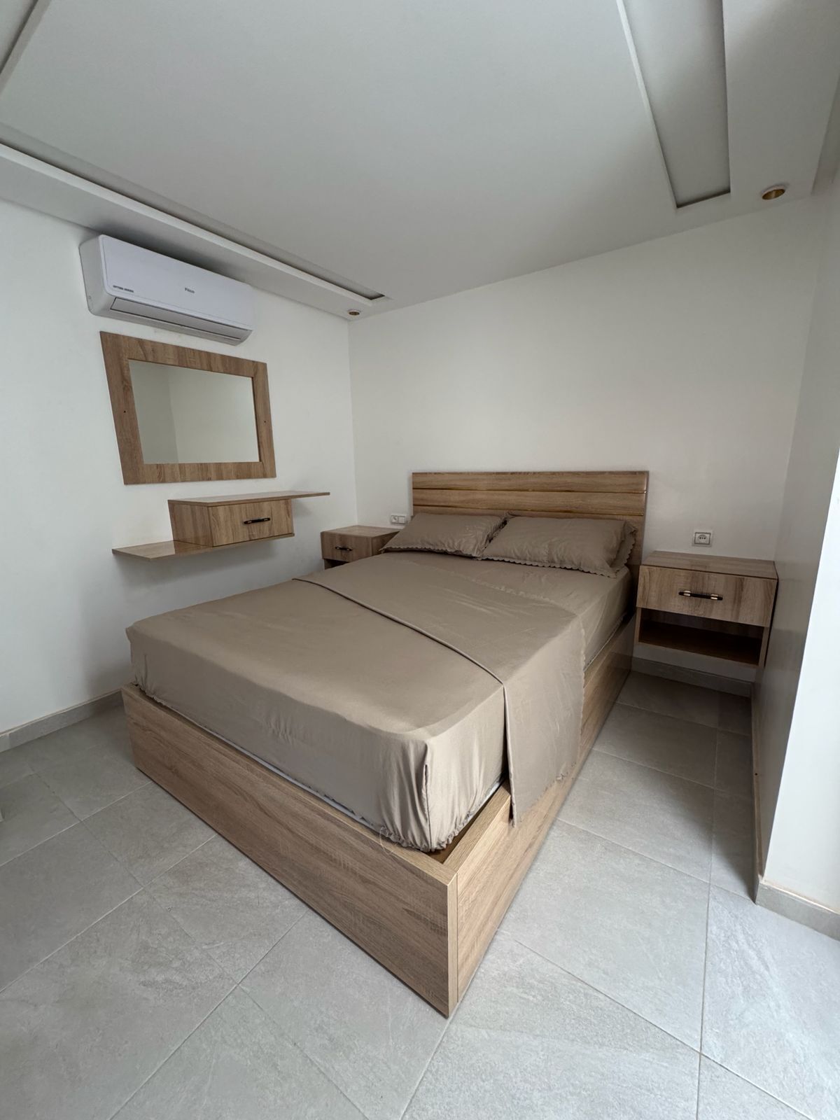 A bedroom with a bed, a nightstand, a bedside table, a mirror, and an air conditioning unit.