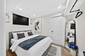A modern bedroom with a bed, a large mirror, a door, and a wall-mounted TV. - Cozy Modern Walkerville
Basement Apartment (Windsor)
