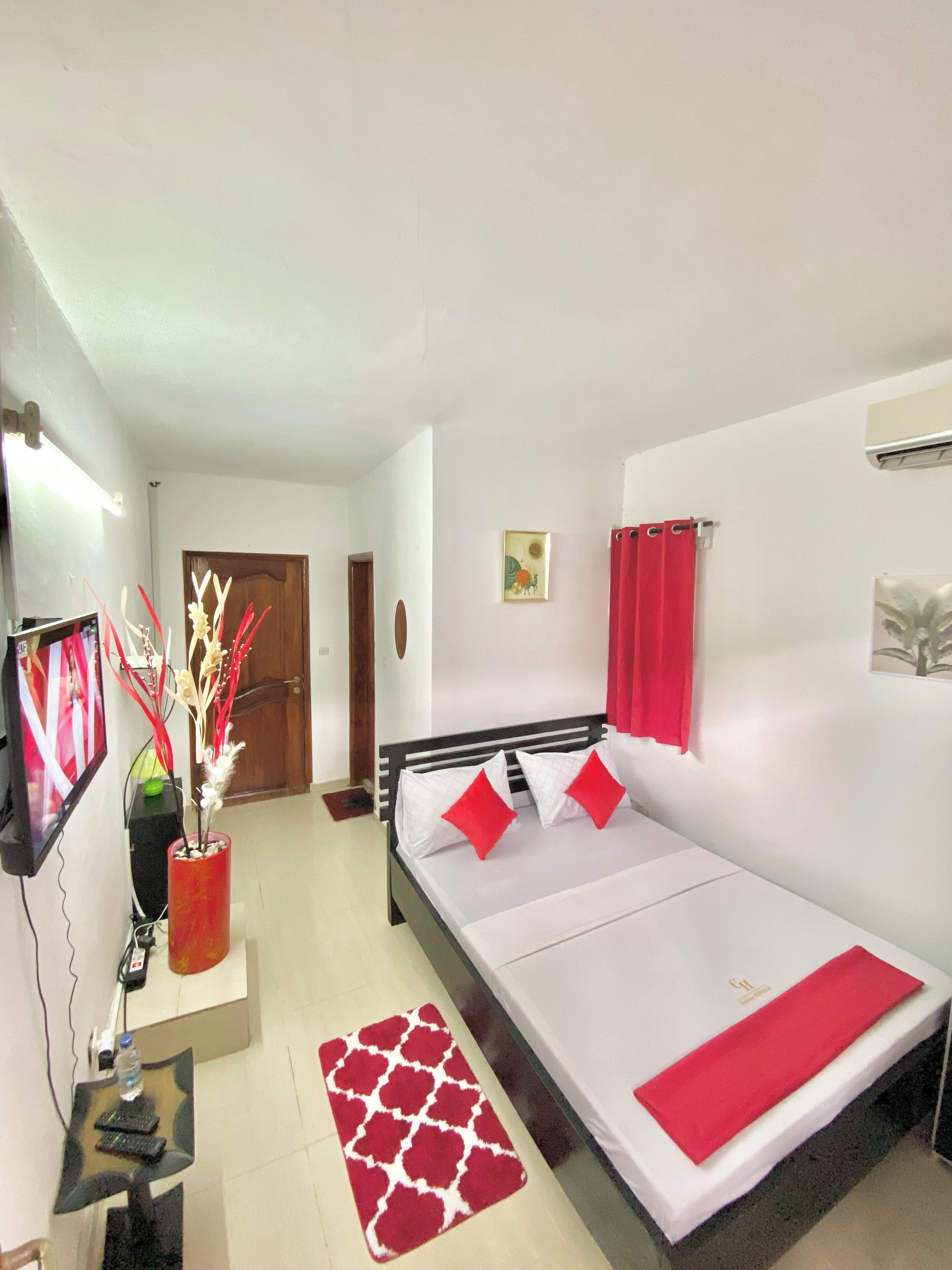 A bedroom with a bed, a TV, a red vase, and a red rug.