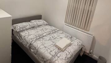 Ein Schlafzimmer mit Bett, Heizkörper, Fenster mit Jalousien, einer Pendelleuchte und einem kleinen Bild an der Wand.