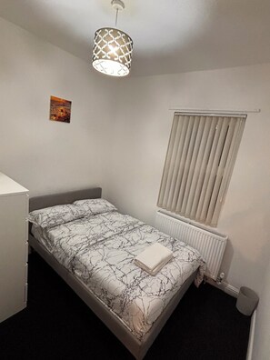 A bedroom with a bed, a radiator, a window with blinds, a hanging light fixture, and a small painting on the wall. - Spacious 4BR Home | Sleeps 7 | 2 Baths | Long Stays (Manchester)