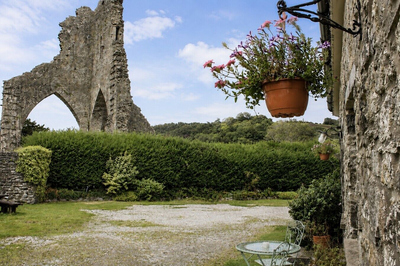 An outdoor area with a stone archway, a hanging flower pot, a table with chairs, and a hedge.