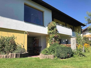 Ein zweistöckiges Haus mit Balkon, großem Fenster und Garten mit verschiedenen Pflanzen und einer Ziegelmauer.