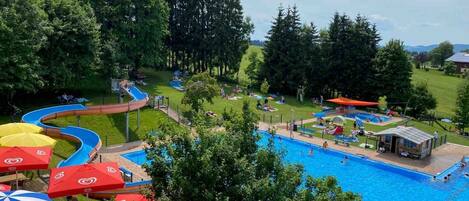 A swimming pool with a water slide, surrounded by trees and umbrellas.