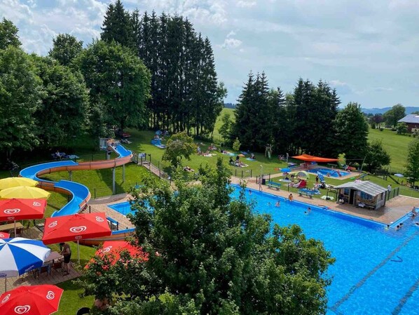 A swimming pool with a water slide, surrounded by trees and umbrellas.