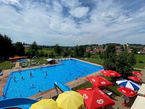 Ein Schwimmbecken mit mehreren Bahnen, umgeben von Sonnenschirmen und Blick auf ein Dorf im Hintergrund.