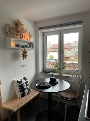 A dining area with a round table, a bench, a chair, a window with a view of houses, and wall-mounted shelves with decorative items. - Modern Oasis of Calm in Allgäu with Private Terrace and Wi-Fi (Oberreute)