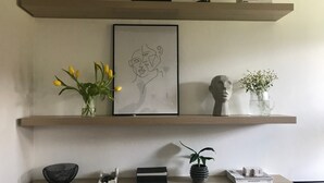 Ein Regal mit einem gerahmten Kunstwerk, einer Vase mit gelben Blumen, einer Büste und einem kleinen Topfpflanzen.