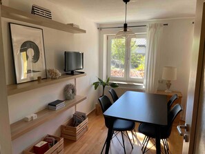 Un comedor moderno con una mesa, sillas, un televisor en un estante y un ventanal con vistas a árboles.