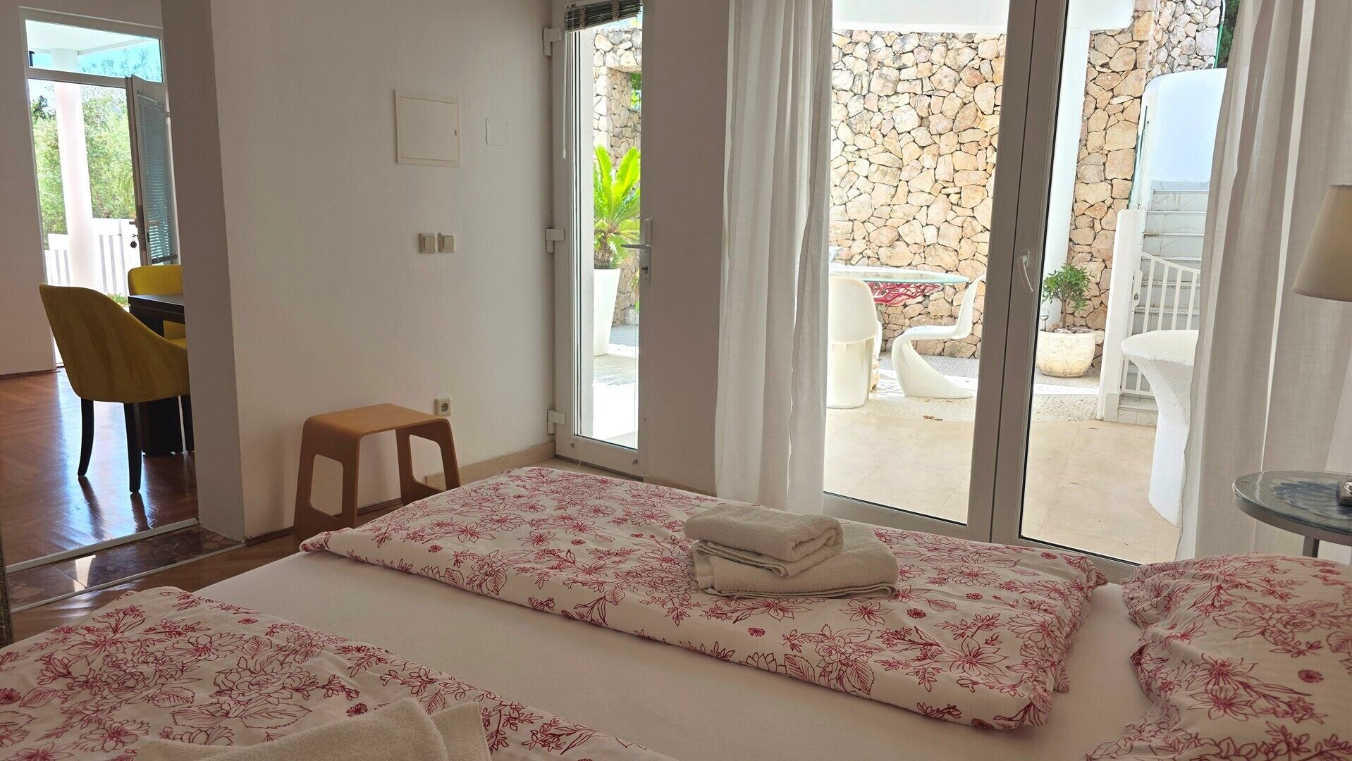 Una habitación con una cama, un taburete, una silla y una mesa. Se aprecia vista de un espacio exterior con un muro de piedra y plantas.
