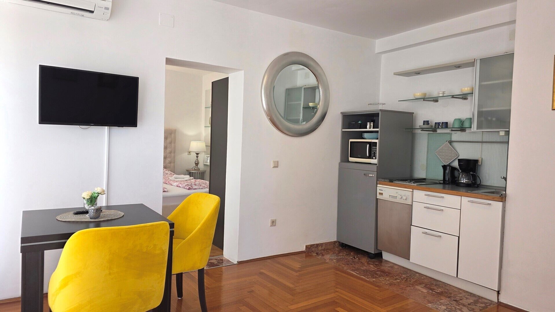 Habitació d'hotel senzilla amb televisió, una petita zona de menjador amb cadires grogues, cuina amb microones i un mirall rodó.