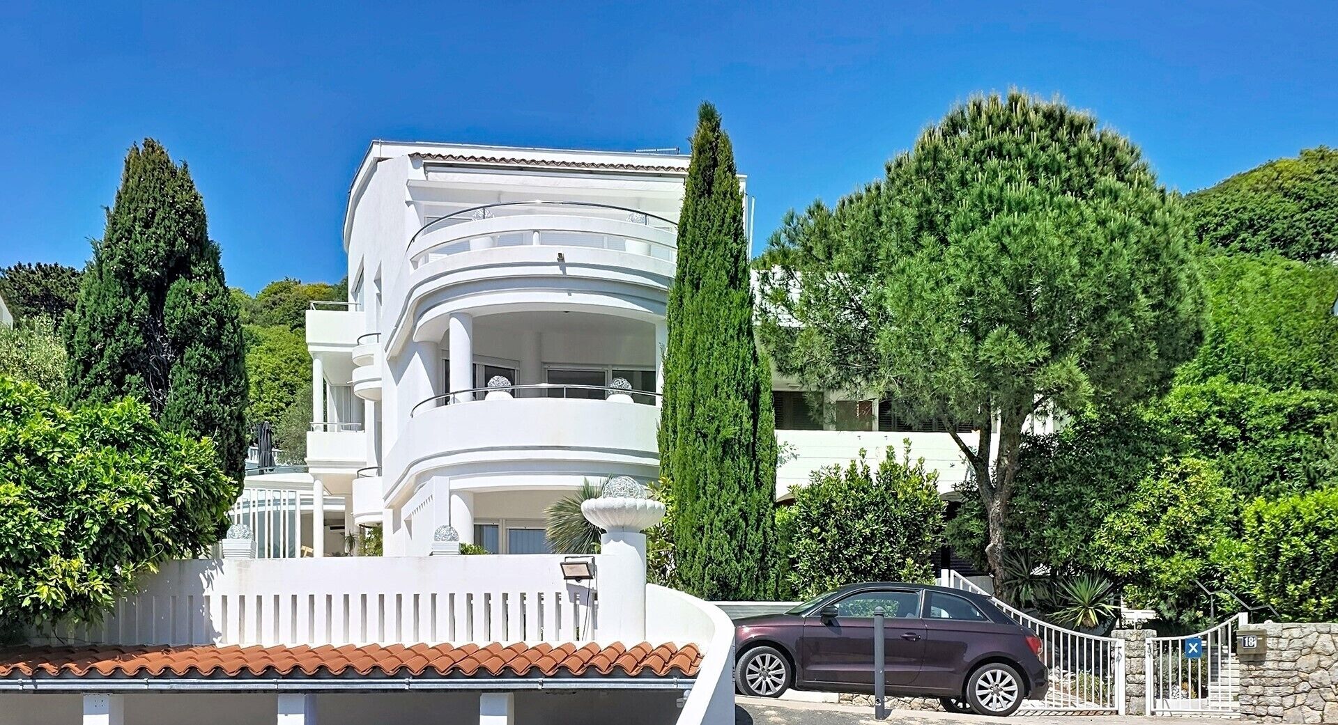 Un immeuble blanc à plusieurs étages, doté de balcons, entouré d’arbres et avec une voiture garée à proximité.