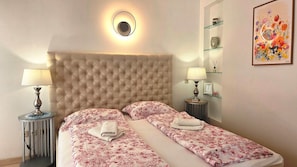 Ein Hotelzimmer mit einem gesteppten Kopfteil, zwei Nachttischen mit Lampen, einer blumigen Tagesdecke und einem gerahmten Blumenbild an der Wand.