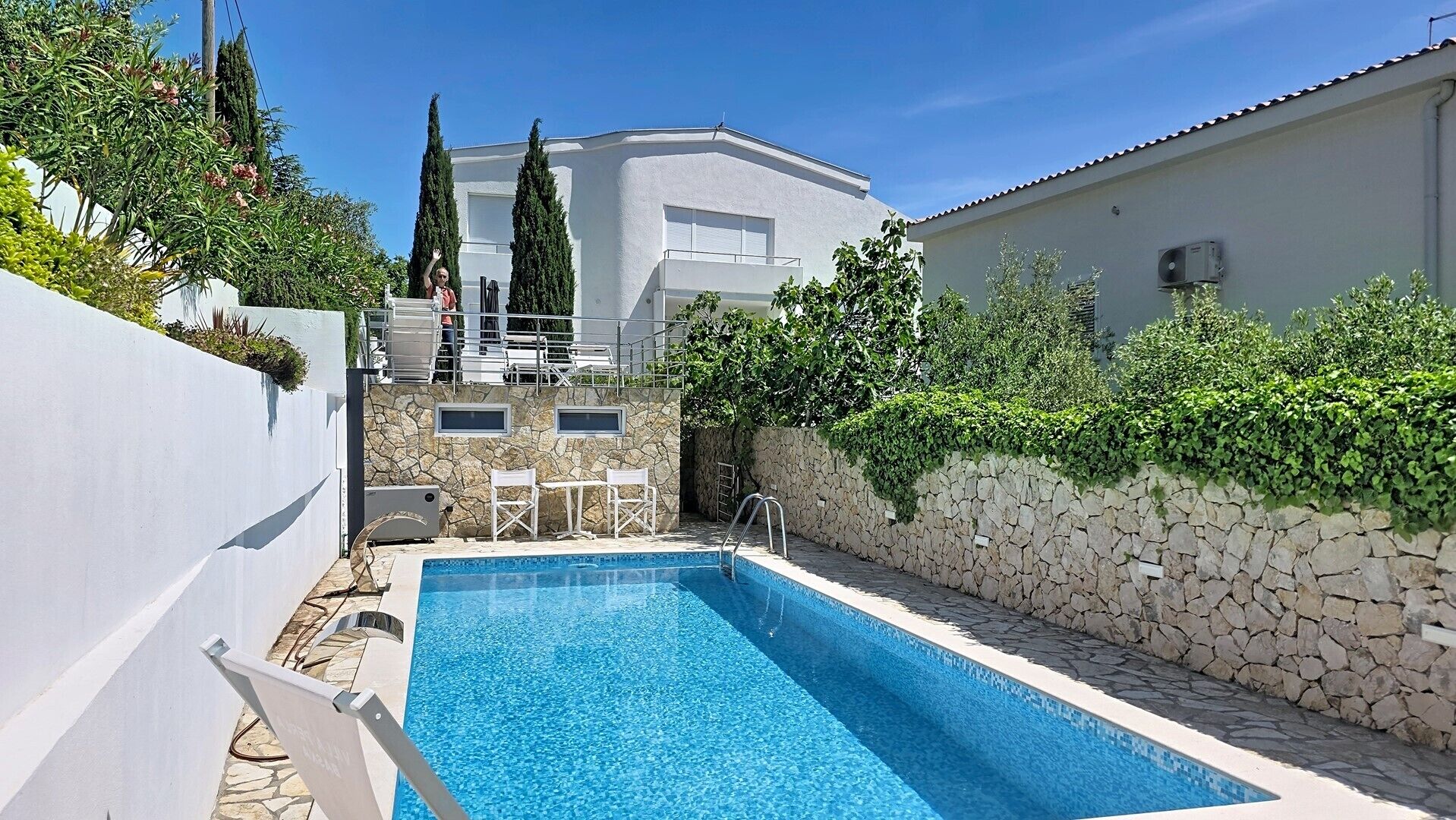 Une piscine aux murs en pierre, un bâtiment blanc et de la verdure en arrière-plan.