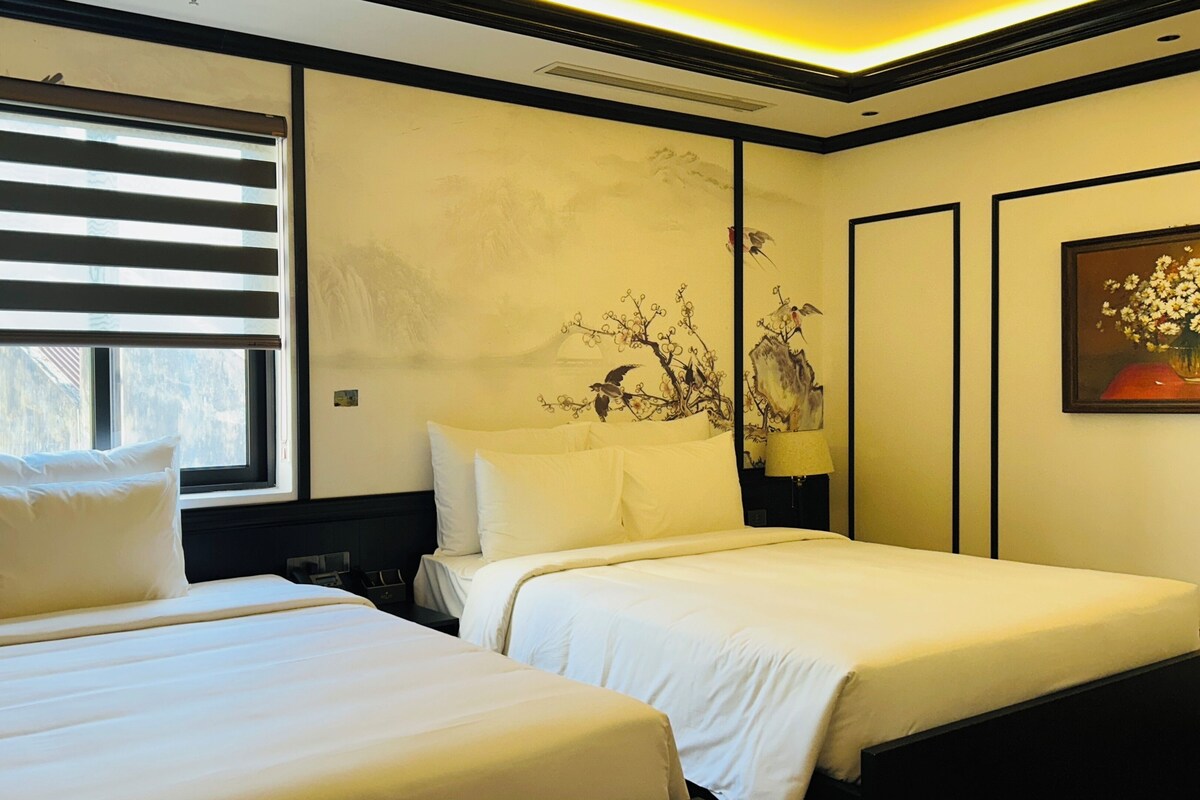 A hotel room with two beds, a large window with blinds, a wall with a floral mural, and a framed painting on the wall.