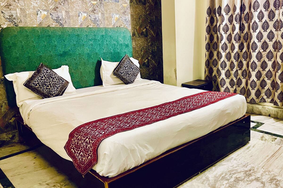 A bed with a red patterned bedspread, two pillows with black and white patterns, a green headboard, and a patterned curtain.