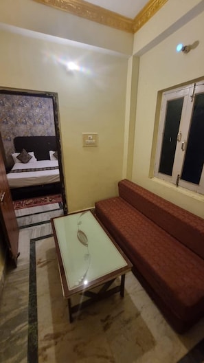 A room with a bed, a sofa, a small table, a window, and a light switch on the wall. - Hotel Magadh Empire (Patna)
