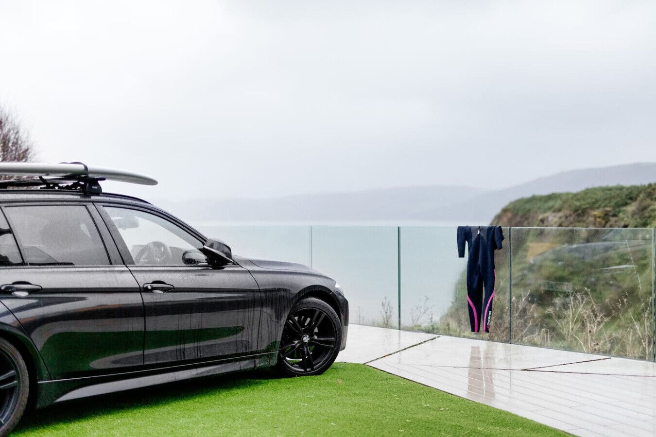 Ein dunkel gefärbtes Auto steht auf einer Rasenfläche mit einer Glasbarriere und Blick aufs Meer.