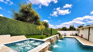 Piscina com borda de pedra e cachoeira, cercada por um hedge bem cuidado e uma área de terraço.