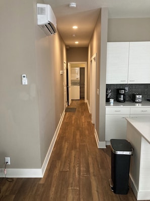 A narrow hallway with a wooden floor leading to a kitchen area with white cabinets and a black trash bin.