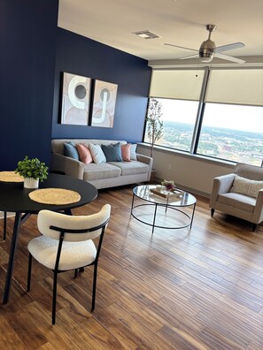 Ein modernes Wohnzimmer mit einer Couch, einem Couchtisch und Sesseln, einem Esstisch mit Untersetzern und einem großen Fenster mit Blick auf die Stadt.