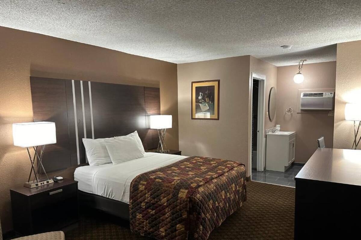 A hotel room with a bed, two bedside lamps, a nightstand, a chair, a desk, a mirror, and a bathroom with a sink and toilet.