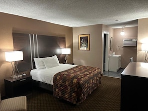 A hotel room with a bed, two bedside lamps, a nightstand, a chair, a desk, a mirror, and a bathroom with a sink and toilet.