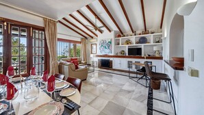 A dining area with a table set for four, a sofa, and a kitchen area with a TV and shelves. - W12 Andalusian style villa with sea views (Mijas Costa)