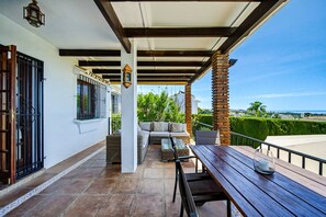 A covered patio with a wooden table and chairs, a sofa, and a view of the ocean. - W12 Andalusian style villa with sea views (Mijas Costa)