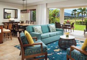 A spacious living room with a sofa, chairs, a dining table, and a view of a lush garden and mountains.