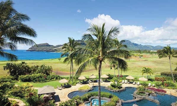 A resort with a pool area, palm trees, and a view of the ocean and mountains.