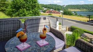 A balcony with wicker furniture, two drinks with garnishes, and a scenic view of houses and a lake. - Apartment "Seeidyll" for 2–4 Guests (48 m²) in Schalkenmehren by the Lake (Schalkenmehren)