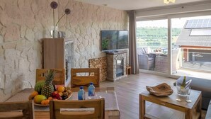 A room with a stone wall, a television, a fireplace, and a balcony with solar panels. - Apartment "Seeidyll" for 2–4 Guests (48 m²) in Schalkenmehren by the Lake (Schalkenmehren)