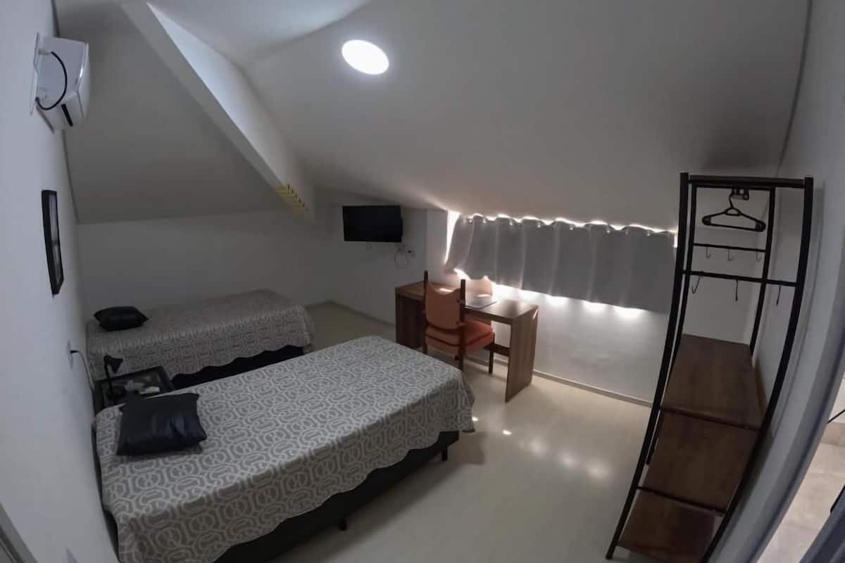 A small, compact room with two single beds, a desk with a chair, a TV mounted on the wall, and a window with curtains.