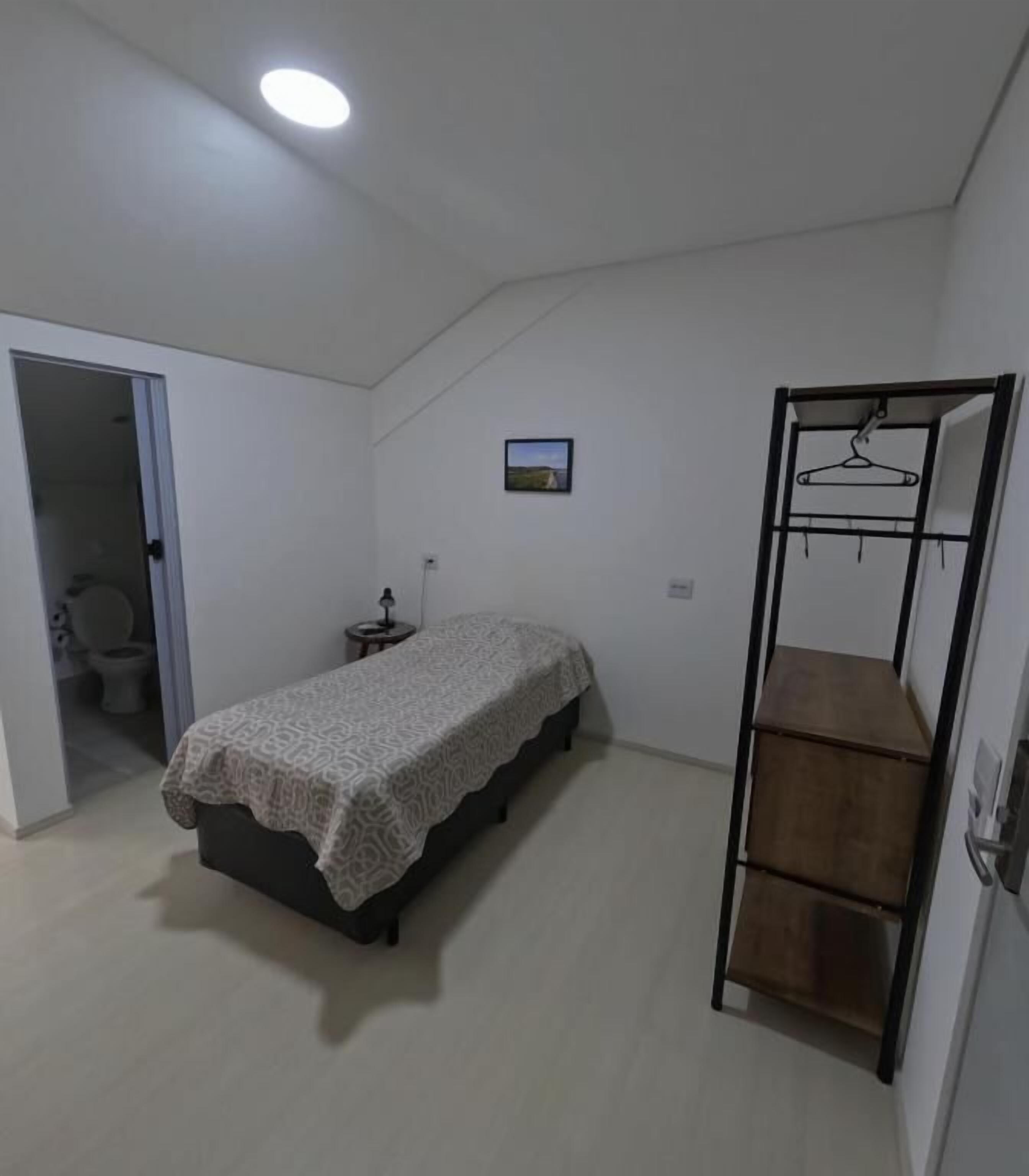 A bedroom with a bed, a hanging shelf, a door leading to a bathroom, and a small bedside table.