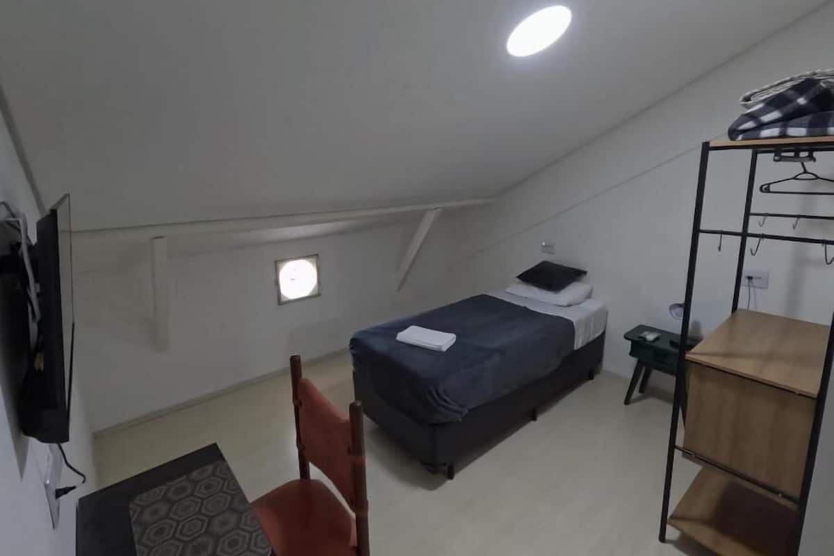 A small, single-room apartment with a bed, a desk, a chair, a TV, a shelf, and a window.