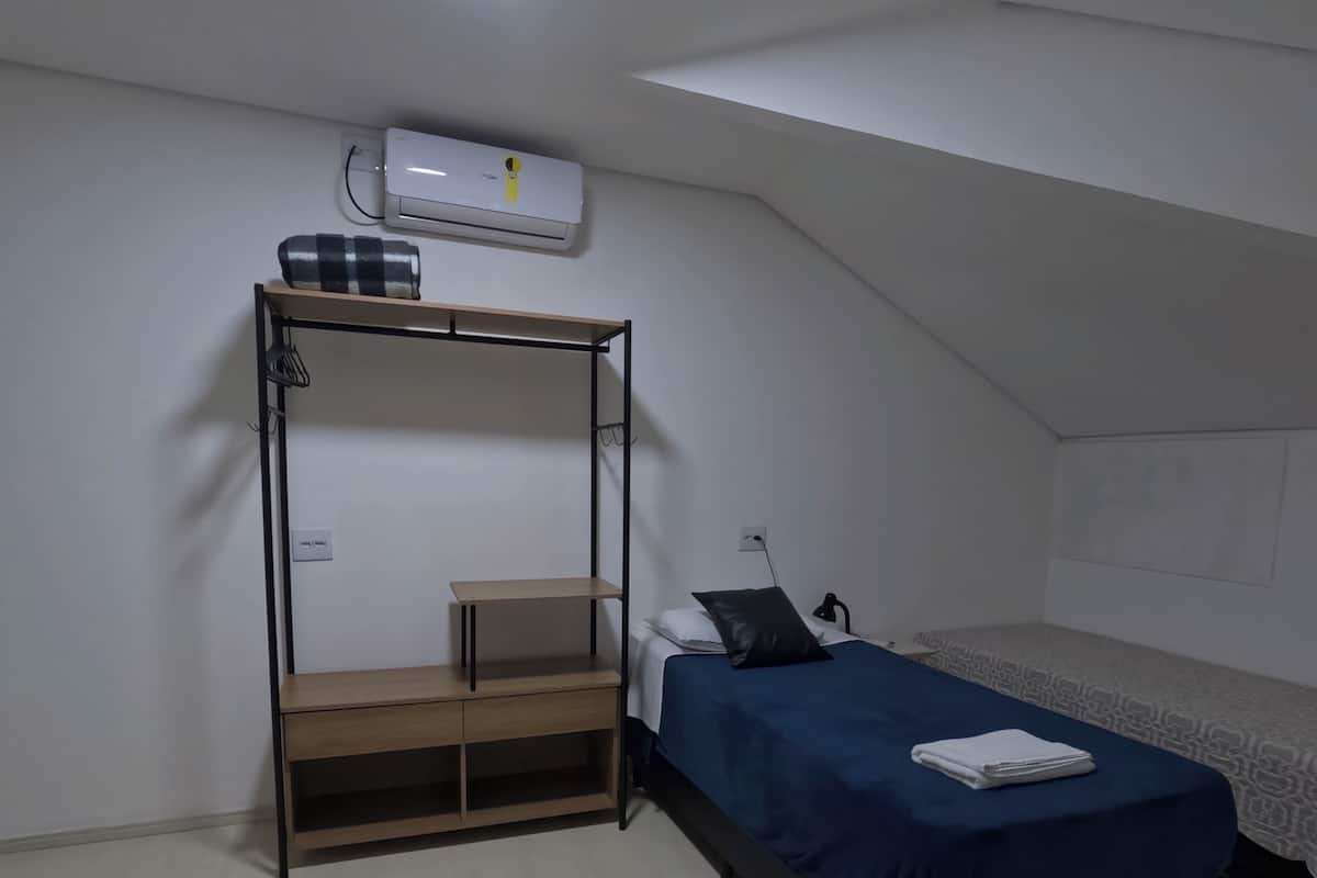 A dorm-style room with a bunk bed, a desk, a chair, a wardrobe, and an air conditioning unit.