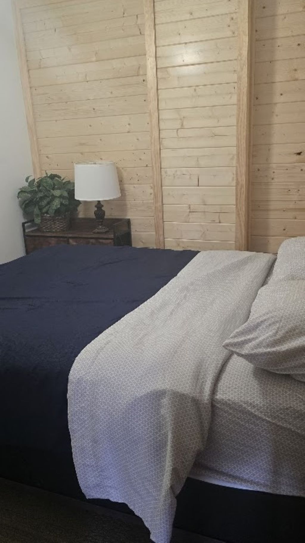Un lit avec une couverture à la fois foncée et claire, une table de chevet avec une lampe, et un mur en bois.