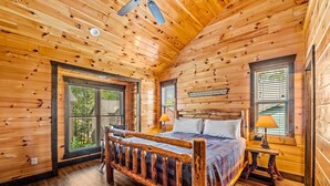 A wooden cabin bedroom with a bed, two windows, a ceiling fan, and a sign on the wall. - The Majestic Pine Retreat (Gatlinburg)