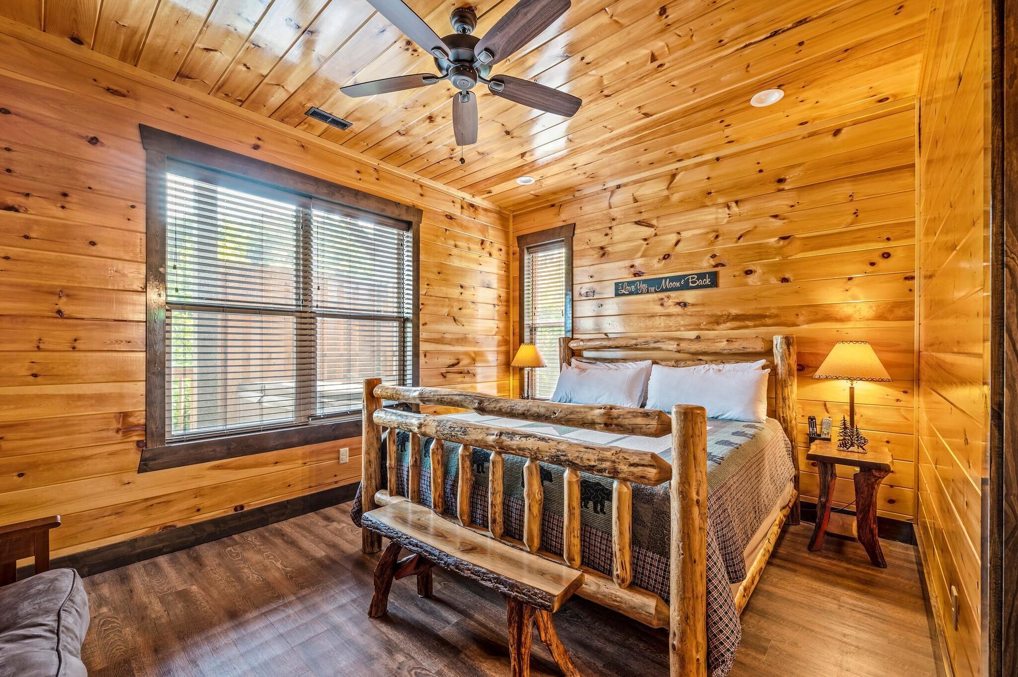 A log cabin bedroom with a wooden bed, a bench, a ceiling fan, and a sign that says 'Love the Moon & Back.'