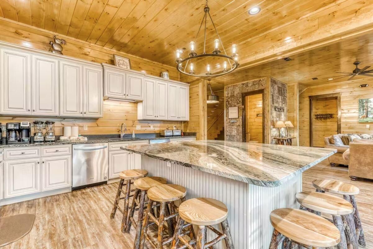 A kitchen with white cabinets, a marble island with stools, and a wooden ceiling.