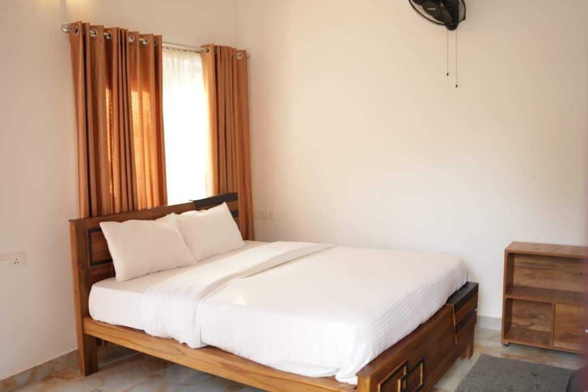 A single bed with a wooden frame and white bedding, a small bedside table, and a wall-mounted fan.
