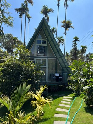 Een huis in een driehoekige vorm met een balkon, omringd door weelderige begroeiing en palmbomen.