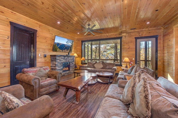A log cabin interior with a fireplace, leather sofas, a wooden coffee table, and a large window with a mountain view. - The Big Poplar Retreat (Gatlinburg)